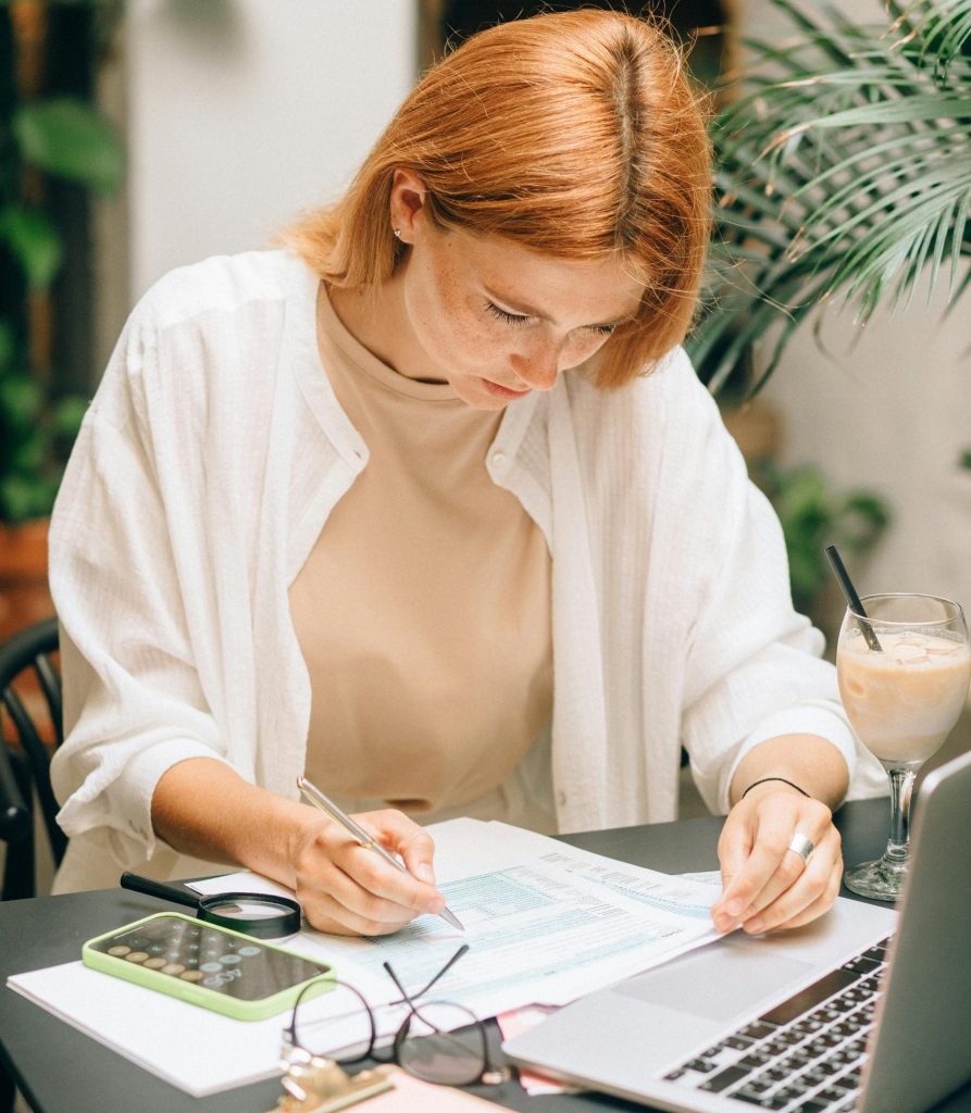 Woman analyzing financial documents using laptop and calculator indoors.